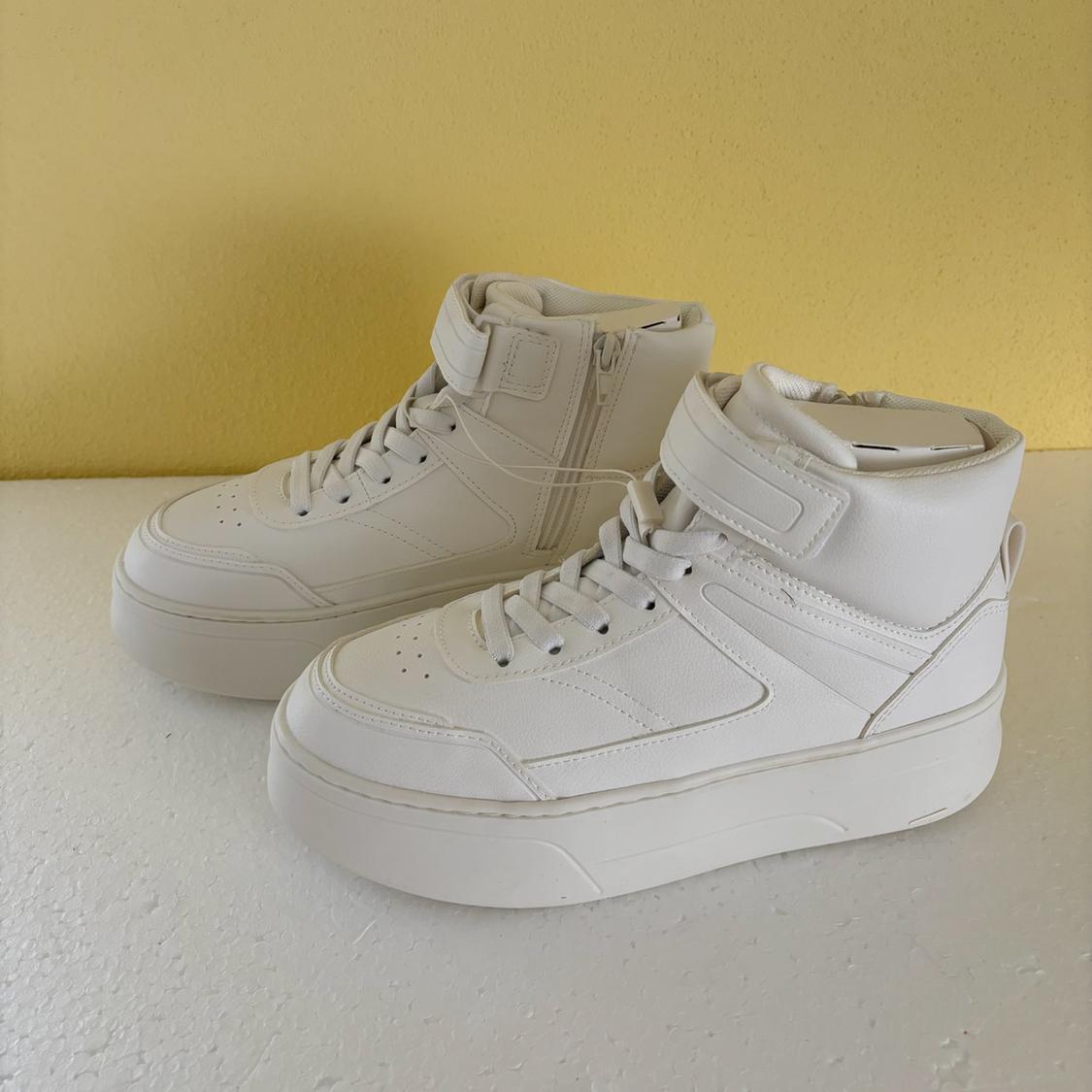 High-top white sneakers with laces and side closure, on a yellow background and white surface.