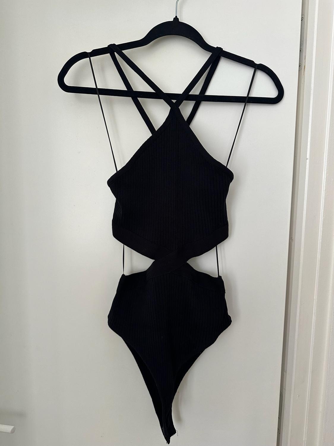Black ribbed halter neck bodysuit with crossover straps and cut-out waist, hanging on a hanger on a white door.