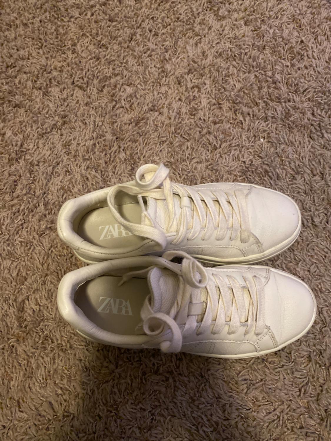 White lace-up trainers on a beige carpet, top view. Interior with "ZARA" logo visible on both trainers.