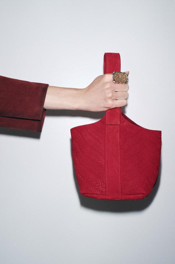Detail červené mini kabelky typu vak s reliéfním tělem s efektem copu a širokým držadlem. Zapínání na klip.