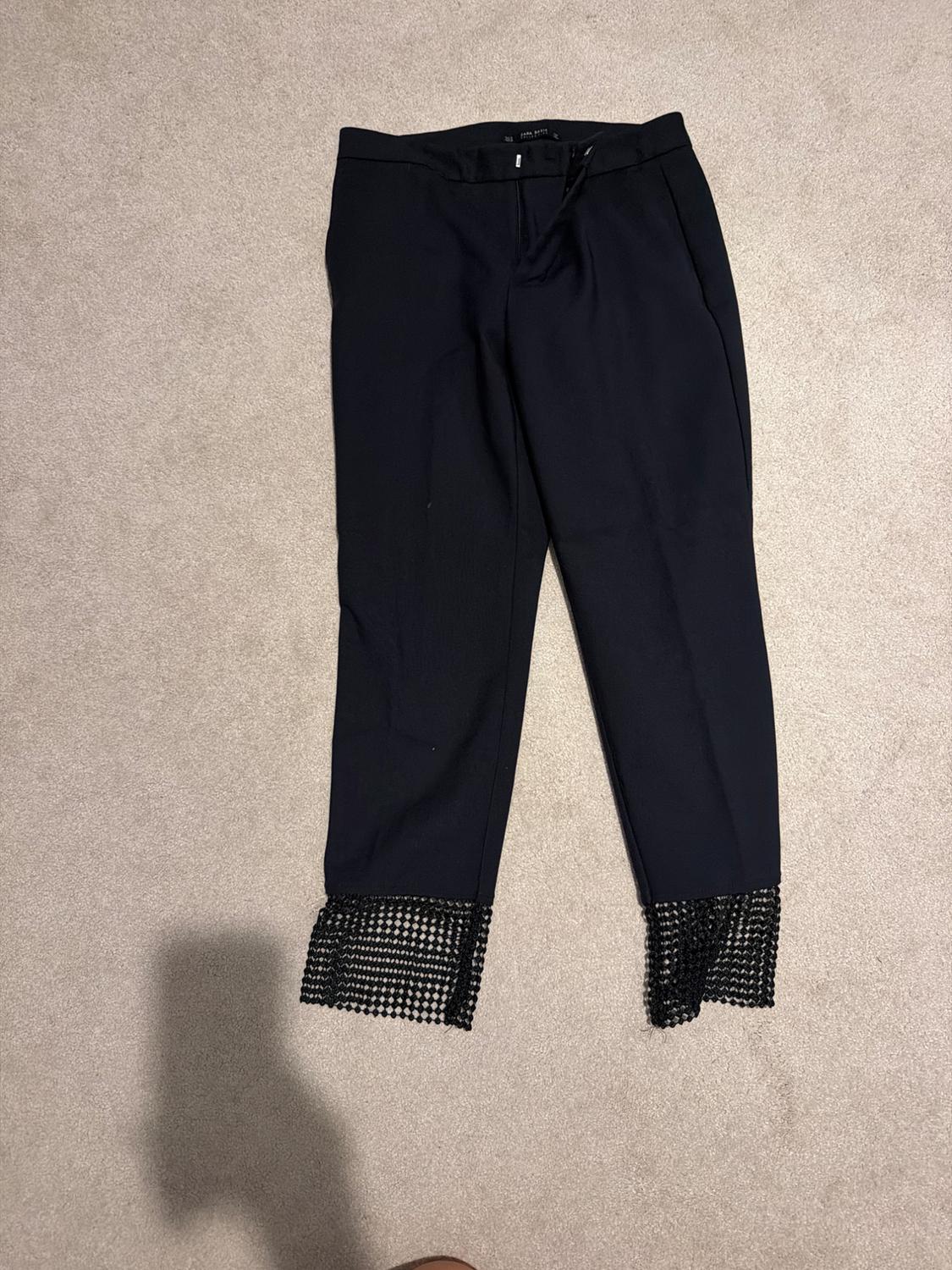 Black trousers laid out on a light-coloured carpet, with a detailed black openwork mesh at the bottom of the legs.