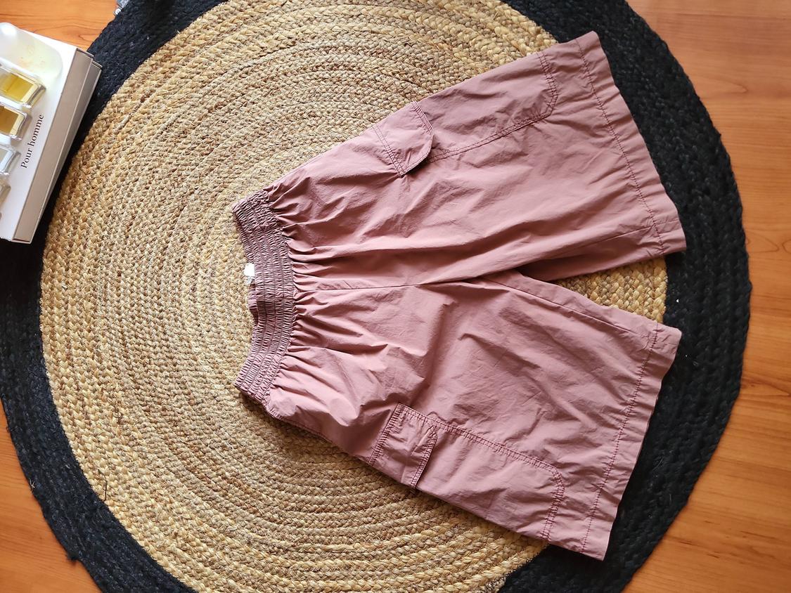 Pink shorts on a round jute rug. To the left, a white box with the text "Pour Homme". Wooden floor visible.