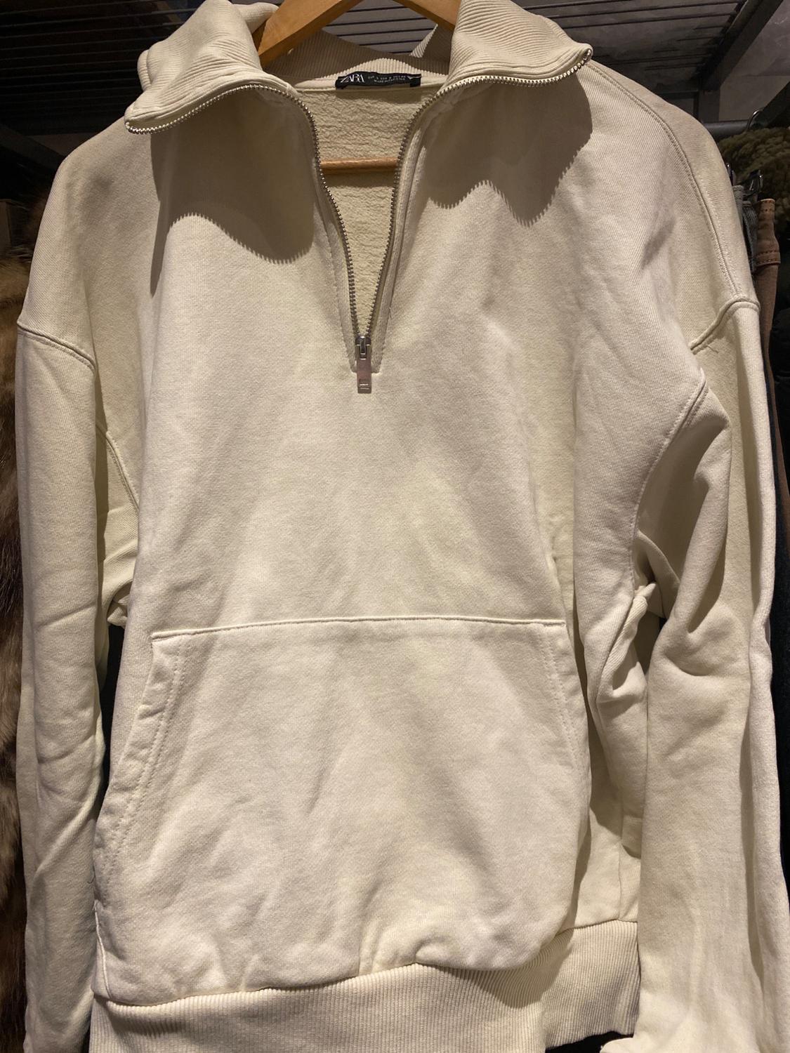 Beige zip-up high-neck sweatshirt hanging on a wooden hanger. Large front pocket visible.