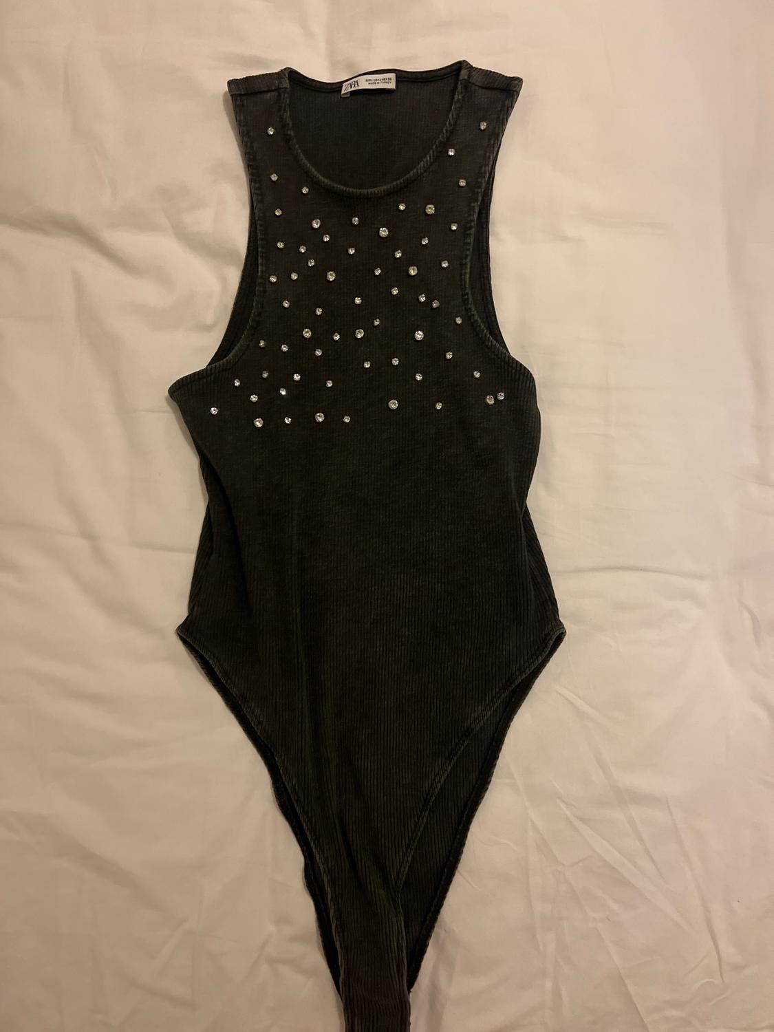 Black sleeveless bodysuit with rhinestone detailing on the front, on a white surface. Visible label on the collar.