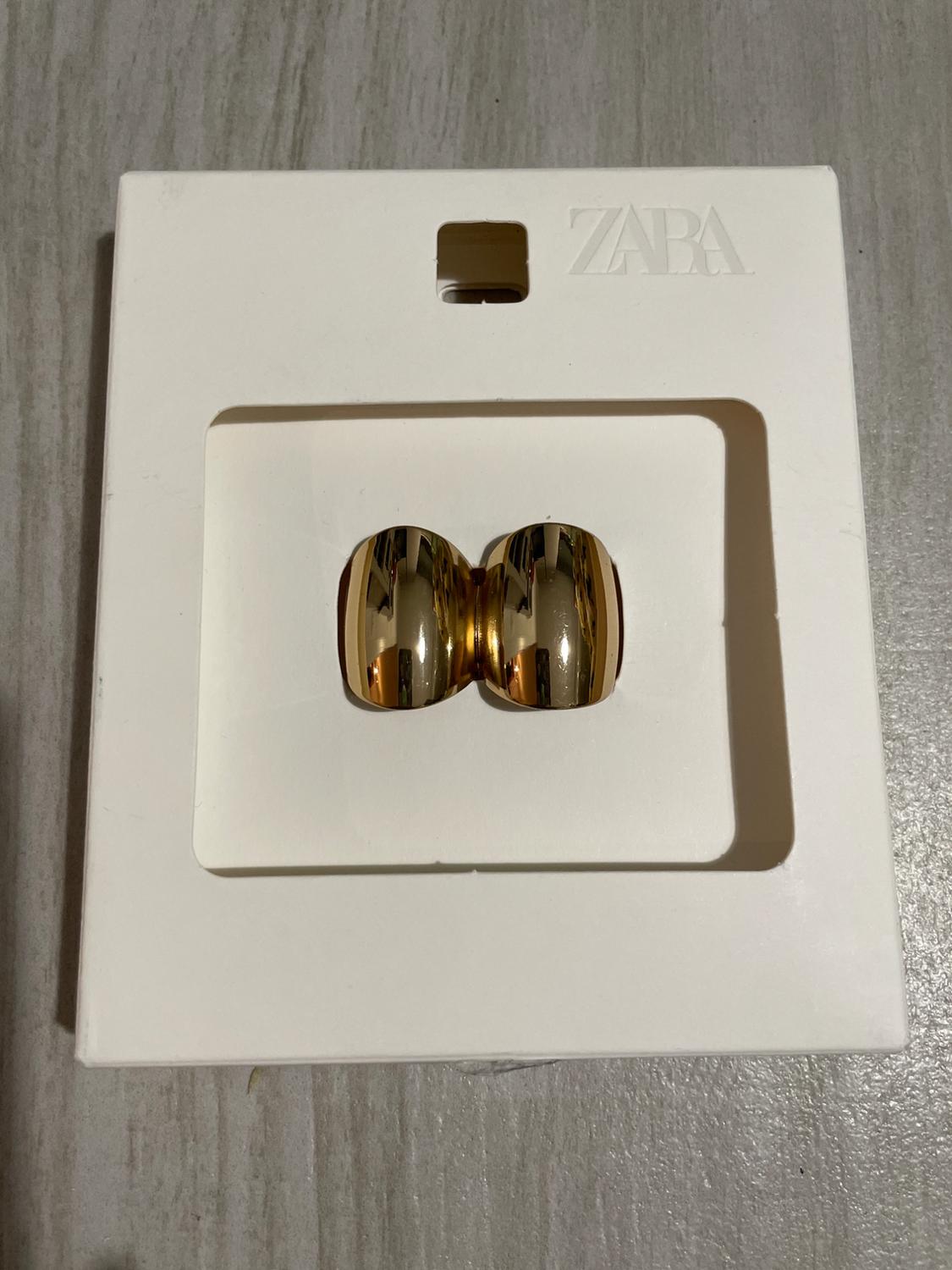 Gold earrings on a white stand with the embossed ZARA logo, on a grey surface.