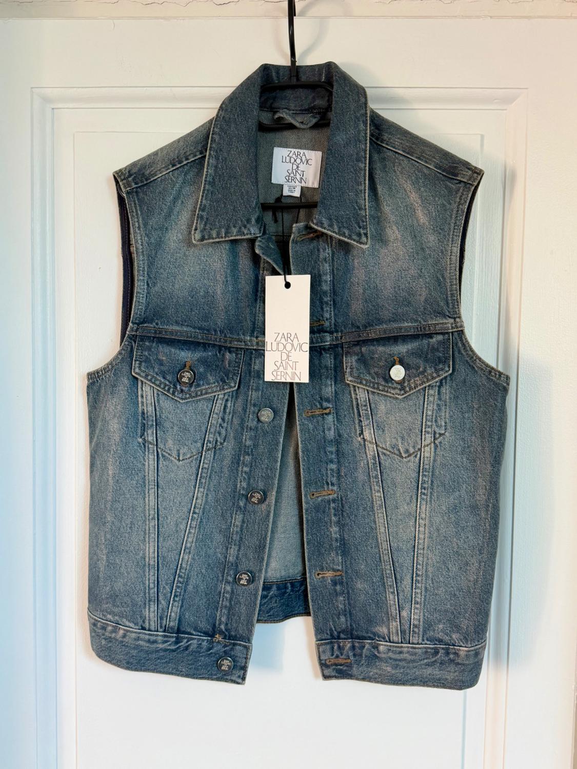 Sleeveless blue denim waistcoat hanging on a hanger. Visible label: "ZARA LUDOVIC DE SAINT SERNIN". White background.