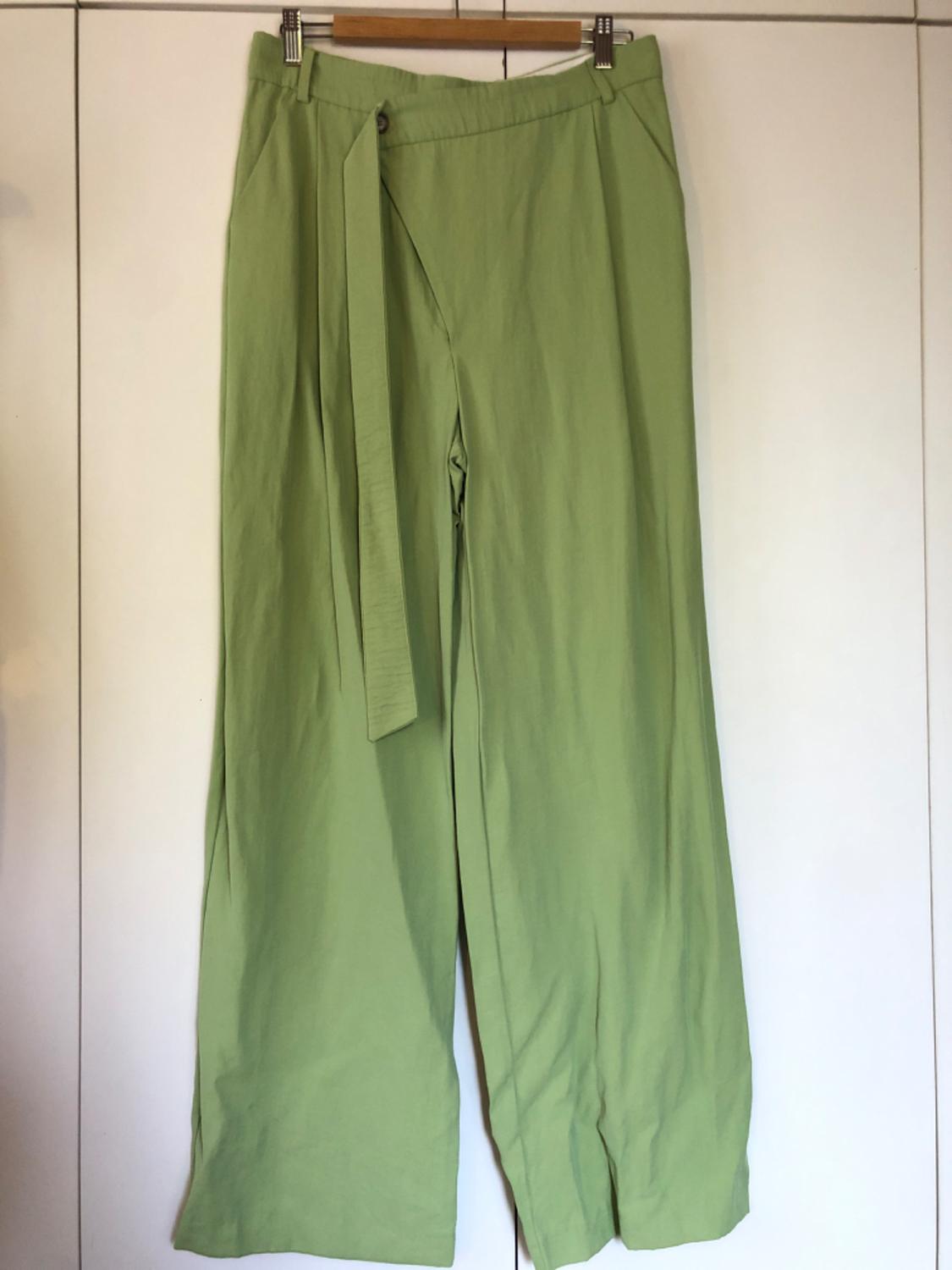 Light green wide-leg trousers with matching belt, hanging on a wooden hanger in front of a white background.