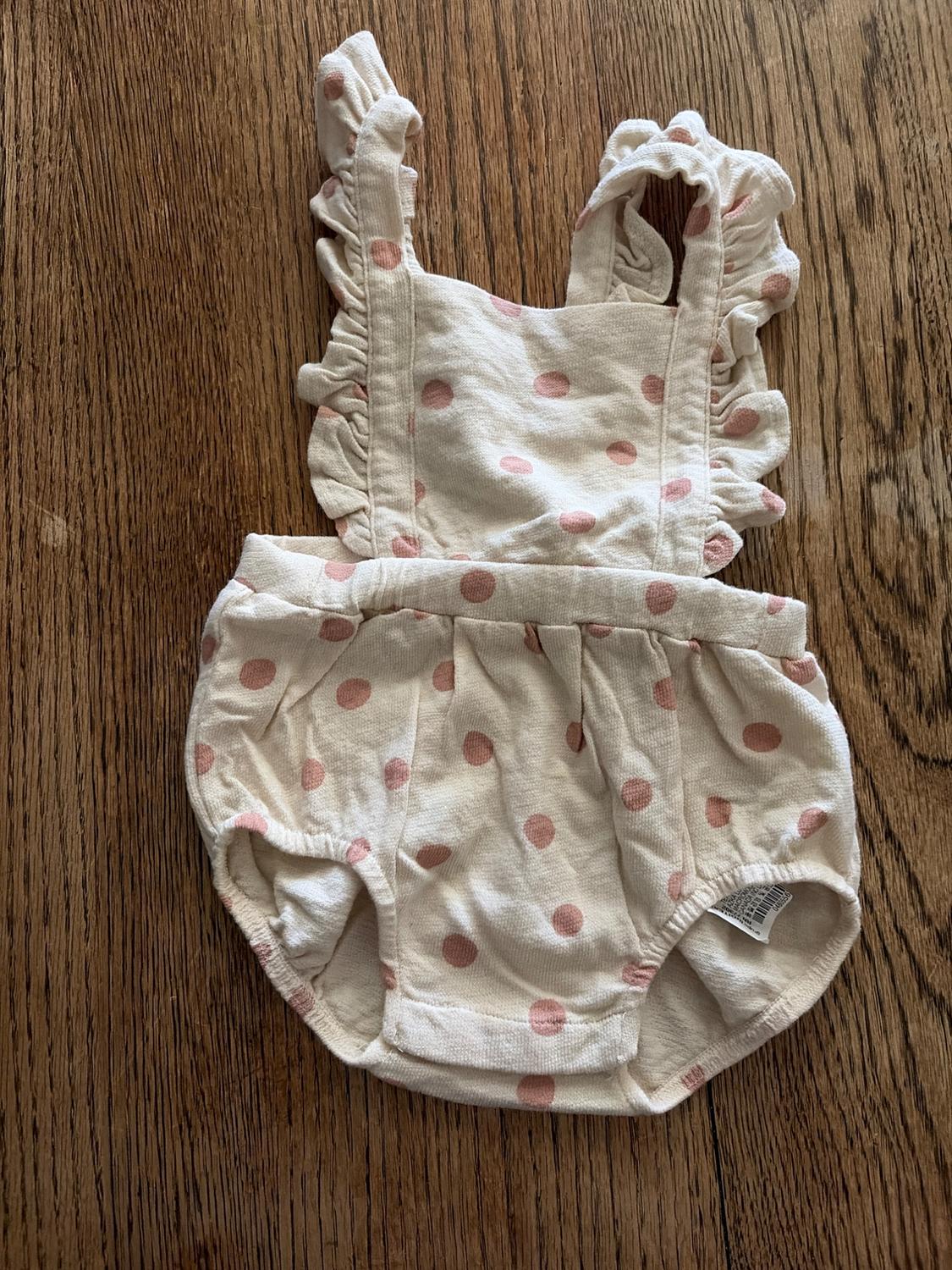 White baby romper with pink polka dots and frills on the straps, on a wooden floor.