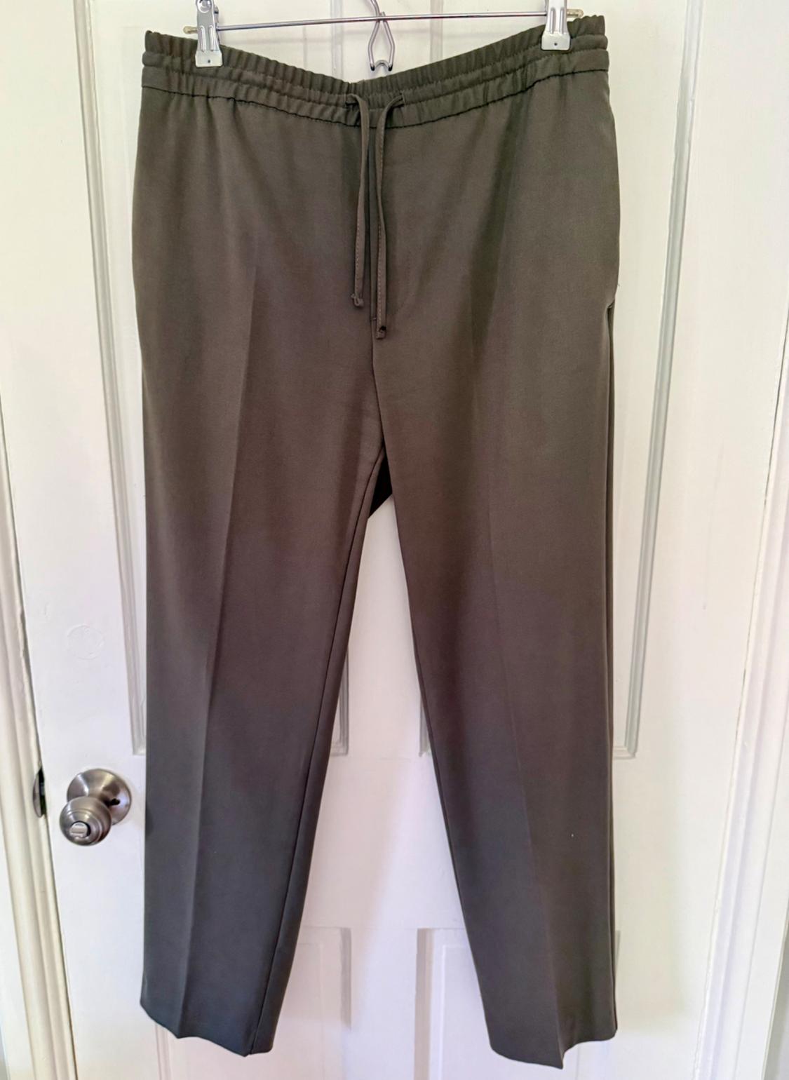 Casual olive green trousers with an elasticated waistband and drawstring, hanging on a hanger over a white door in a relaxed setting.