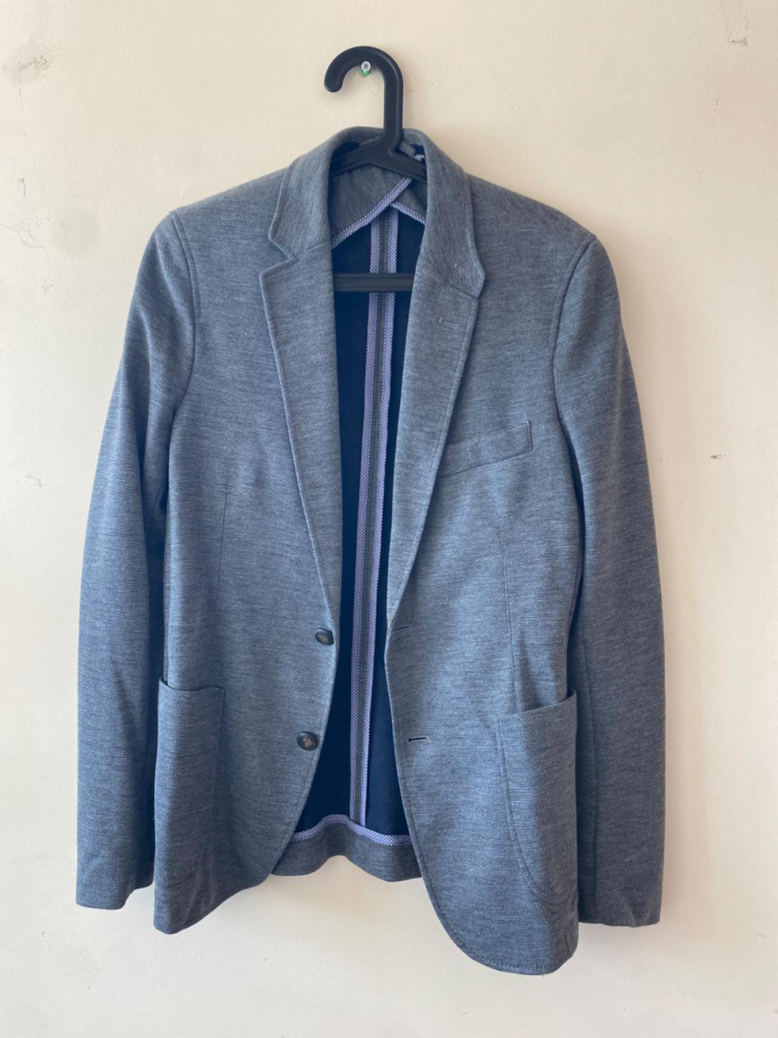 Grey two-button jacket hanging on a black hanger against a white wall. Front pockets visible.