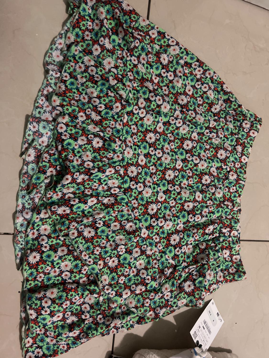 Short skirt with floral print in green, red, and white tones on a black background, placed on a light-coloured floor. Visible tag.