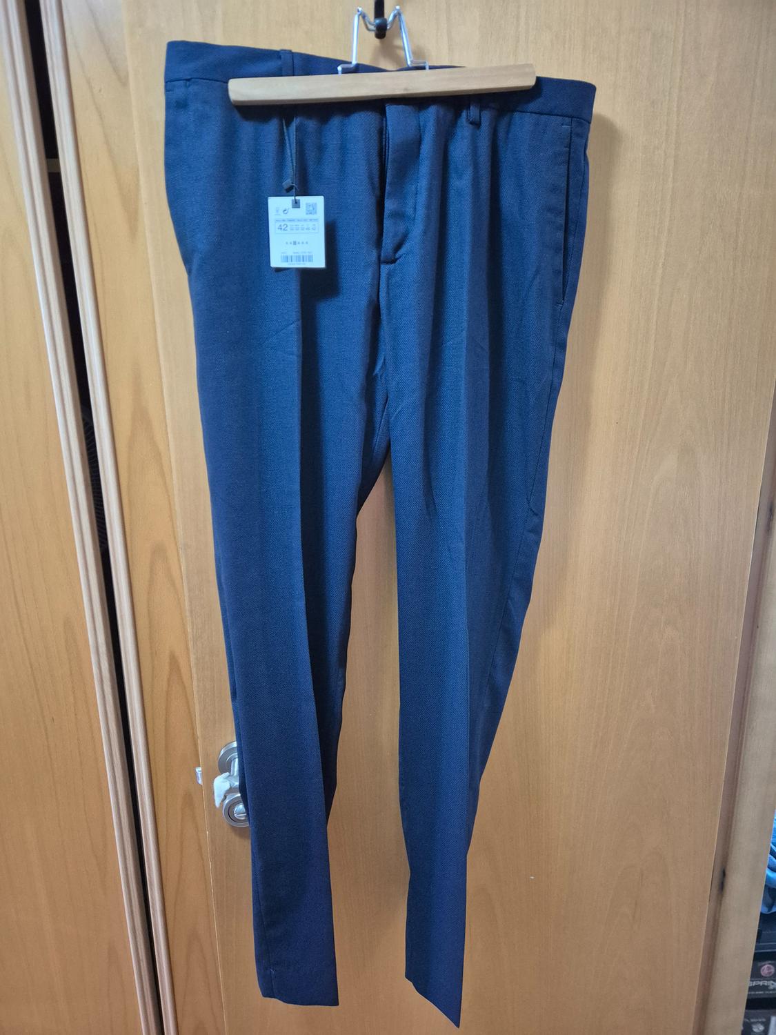 Dark blue trousers hanging on a wooden hanger, with a visible label indicating size 42, against a light wooden door background.