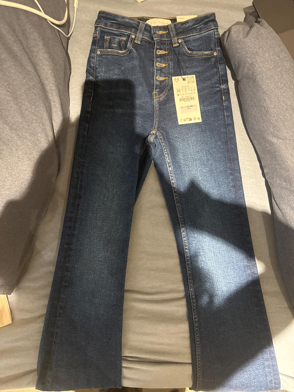 Dark blue jeans with visible buttons and a price tag on a grey bed. Light and shadows in the image.