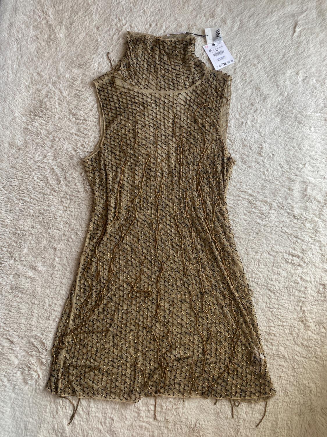 Short, sleeveless, high-neck dress, semi-sheer with black and gold beads, on a fluffy background. Label with ZARA text.