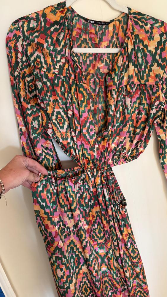 Colourful dress with a geometric print in shades of green, orange, and pink, hanging on a white hanger. Hand adjusting the waist.
