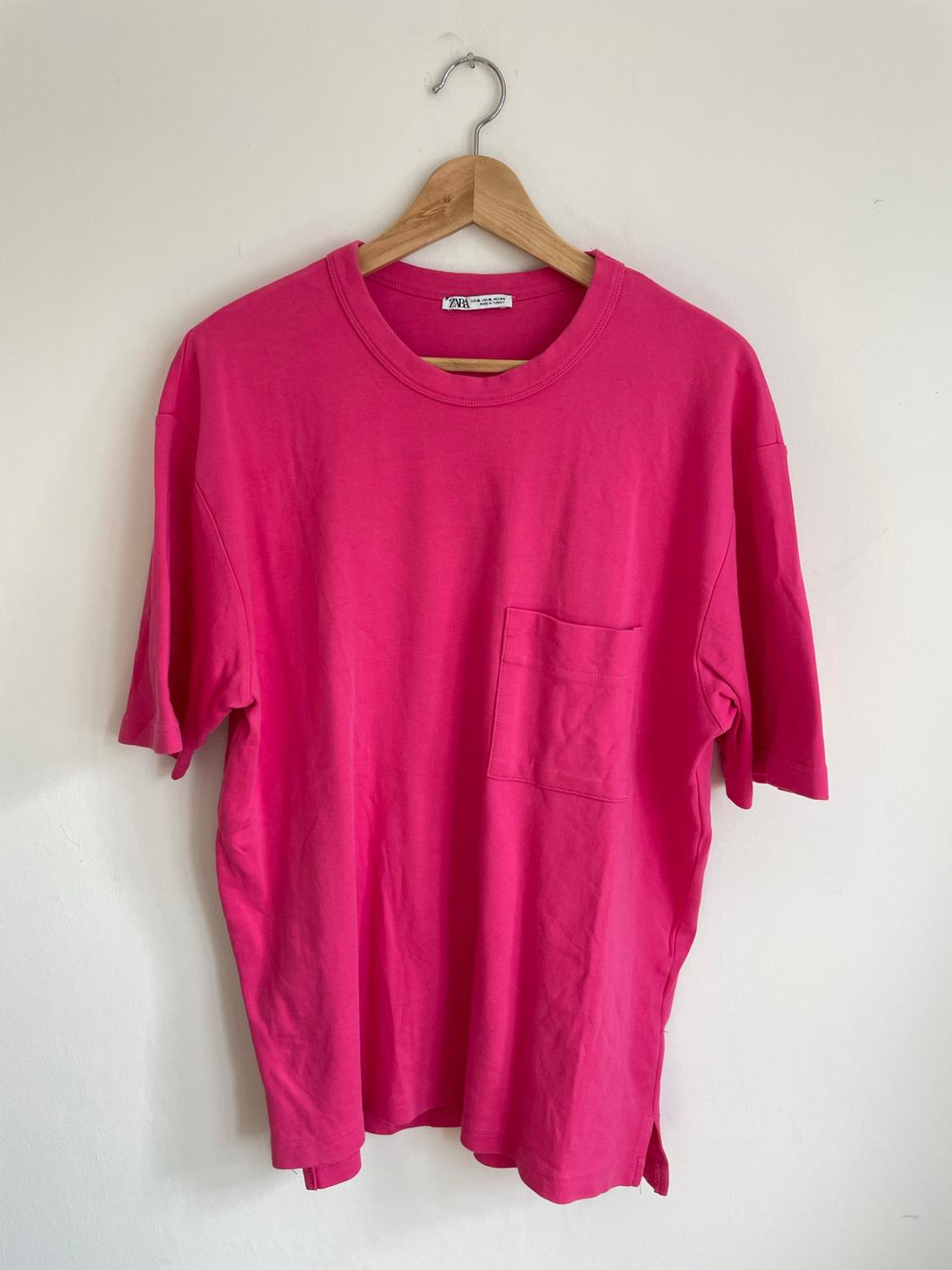 Pink short-sleeved T-shirt with a front pocket, hanging on a wooden hanger against a white wall. Text: ZARA.