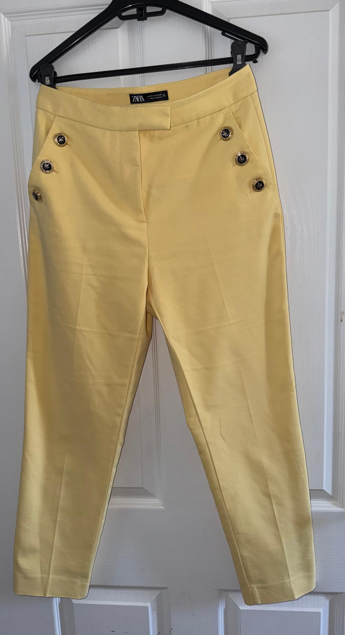 Yellow trousers hanging on a black hanger, with decorative buttons at the top. Visible label: "Zara". White door background.