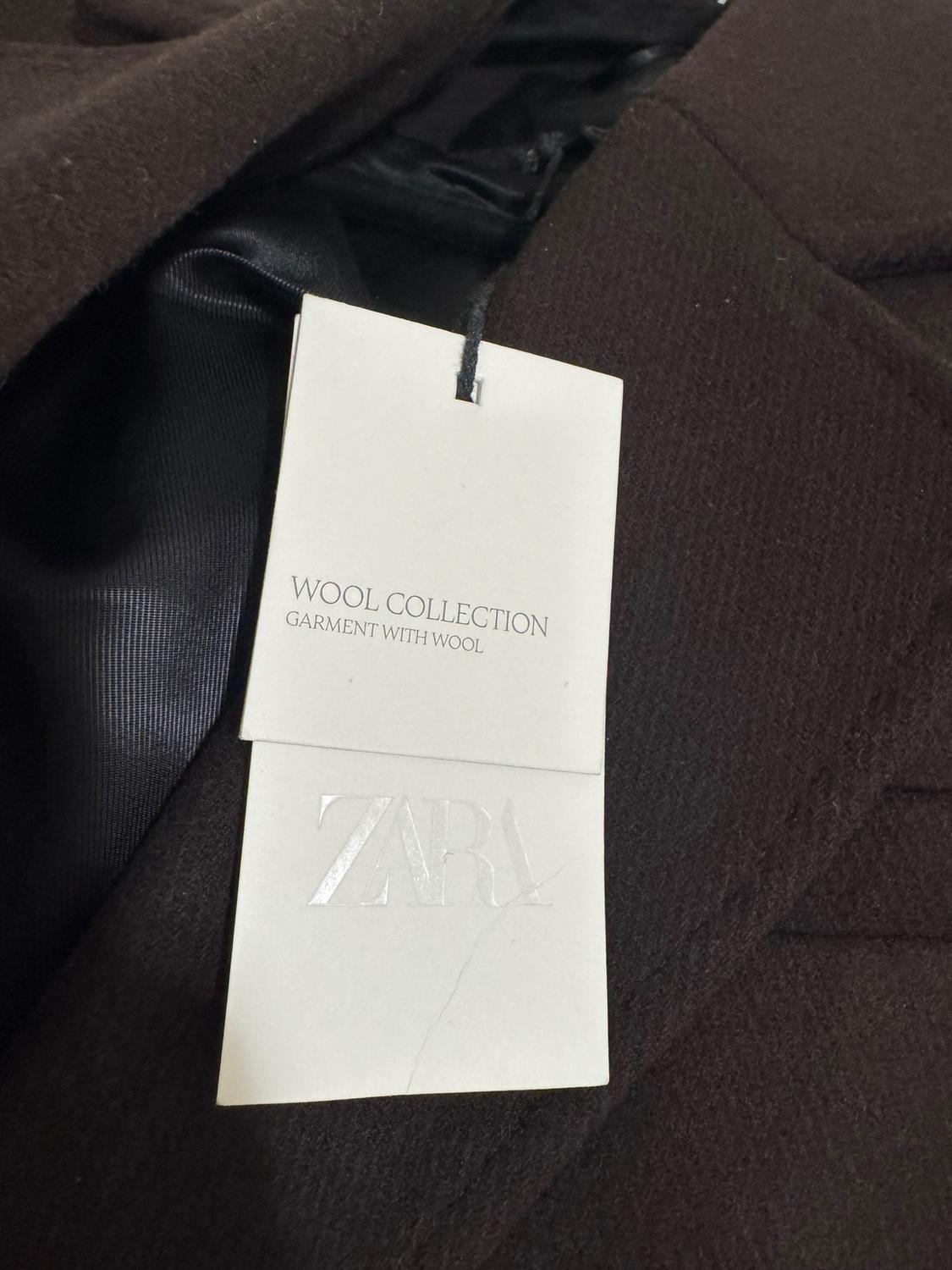 Brown clothing tag with "WOOL COLLECTION GARMENT WITH WOOL" and "ZARA" embossed text. Black fabric visible in the background.