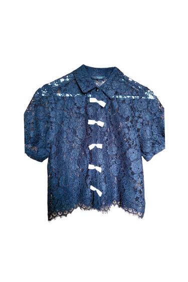 Blue lace blouse with short sleeves and a classic collar. White bow details on the front. White background.