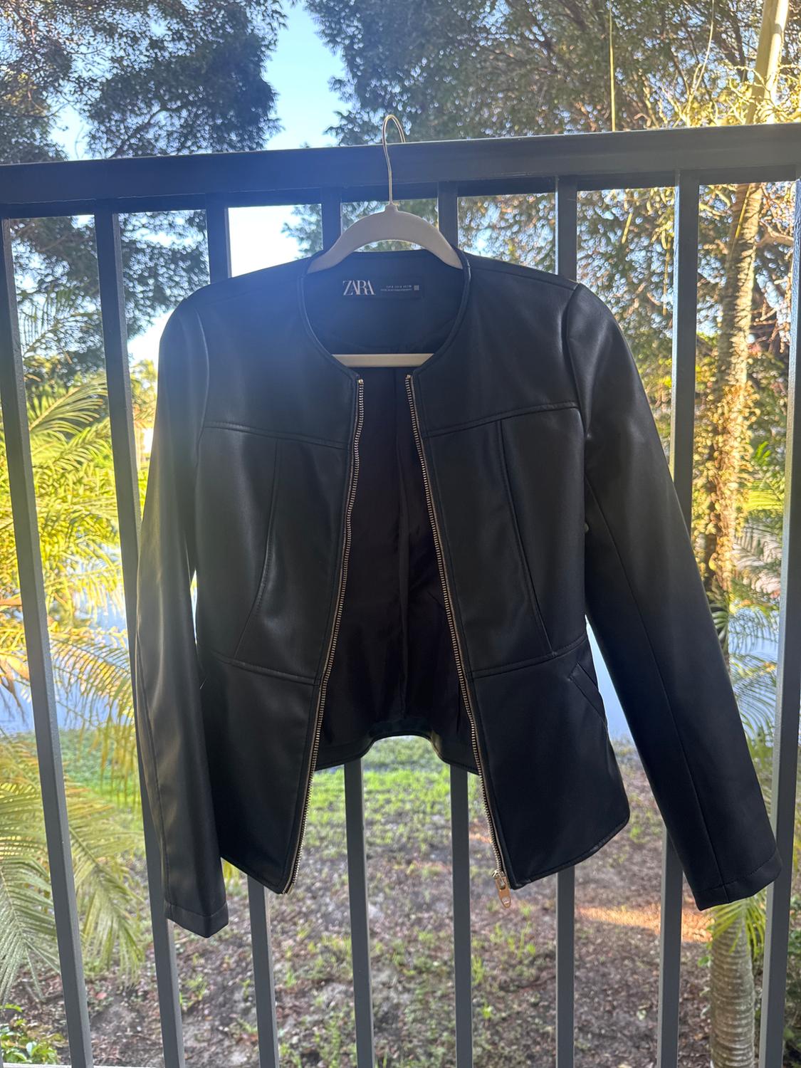 Black jacket hanging on a hanger on a balcony, with trees and blue sky in the background. Visible tag: ZARA.