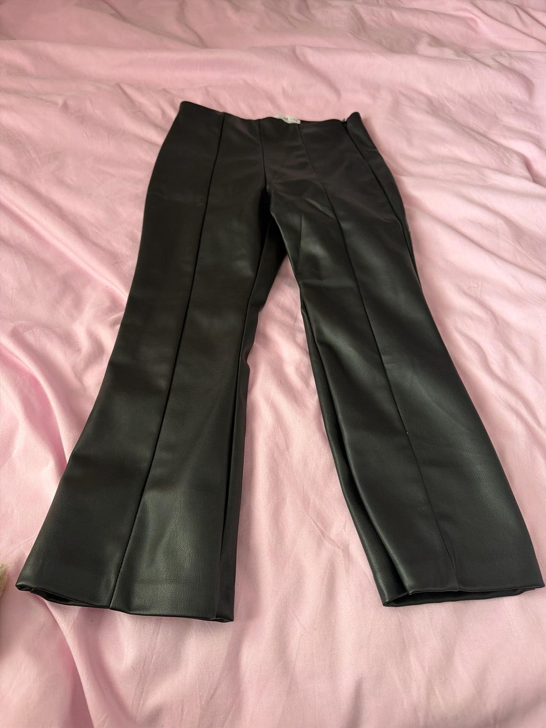 Slightly flared black trousers with front seams, laid out on a crumpled pale pink fabric.