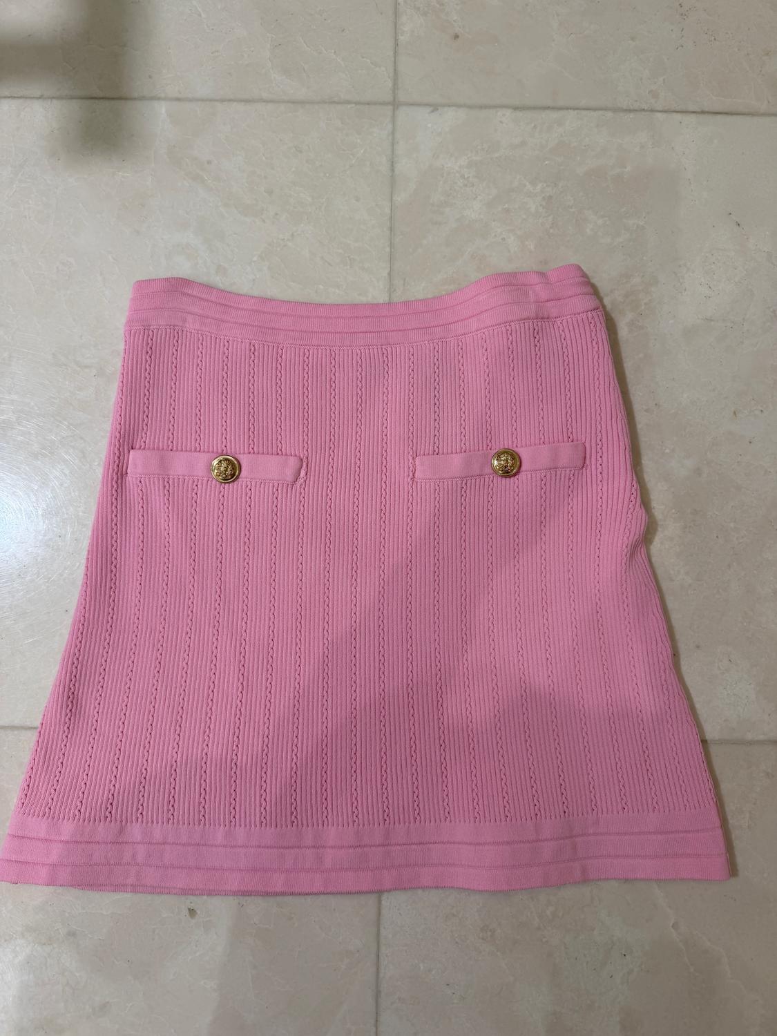 Pink ribbed knit skirt with two decorative gold button pockets, on a light tiled floor.