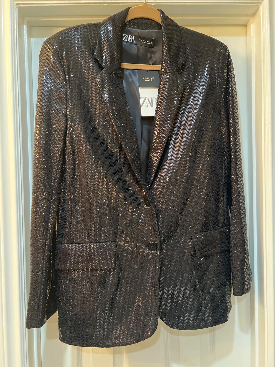 Shiny black jacket hanging on a wooden hanger. Visible label with text: "ZARA" and "FESTIVE DAYS".