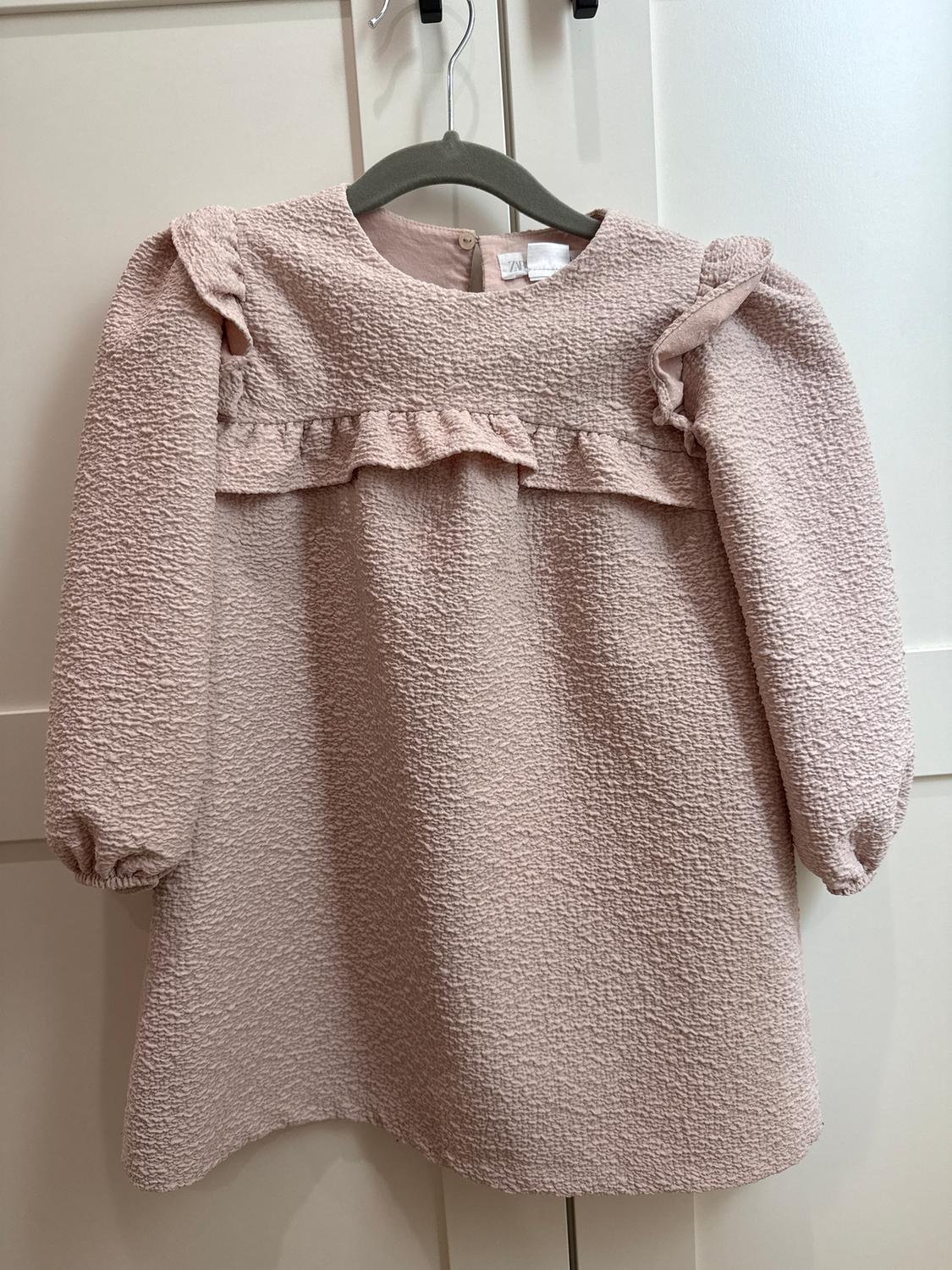 Light pink crinkled texture children's dress with ruffles on the chest, hanging on a grey hanger in front of a white door.