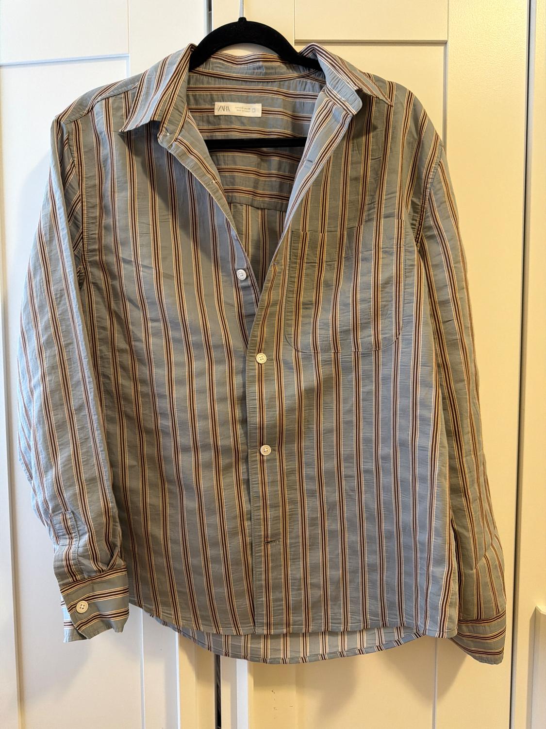 Blue, brown, and white vertical-striped long-sleeved shirt, hanging on a black hanger against a white door. ZARA label visible.