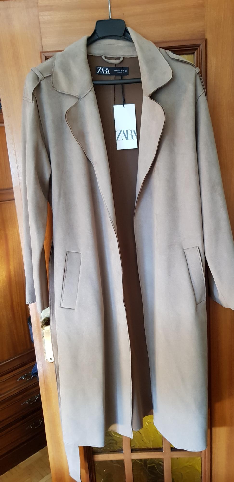 Long beige coat hanging on a wooden door, with a visible Zara label on the collar.