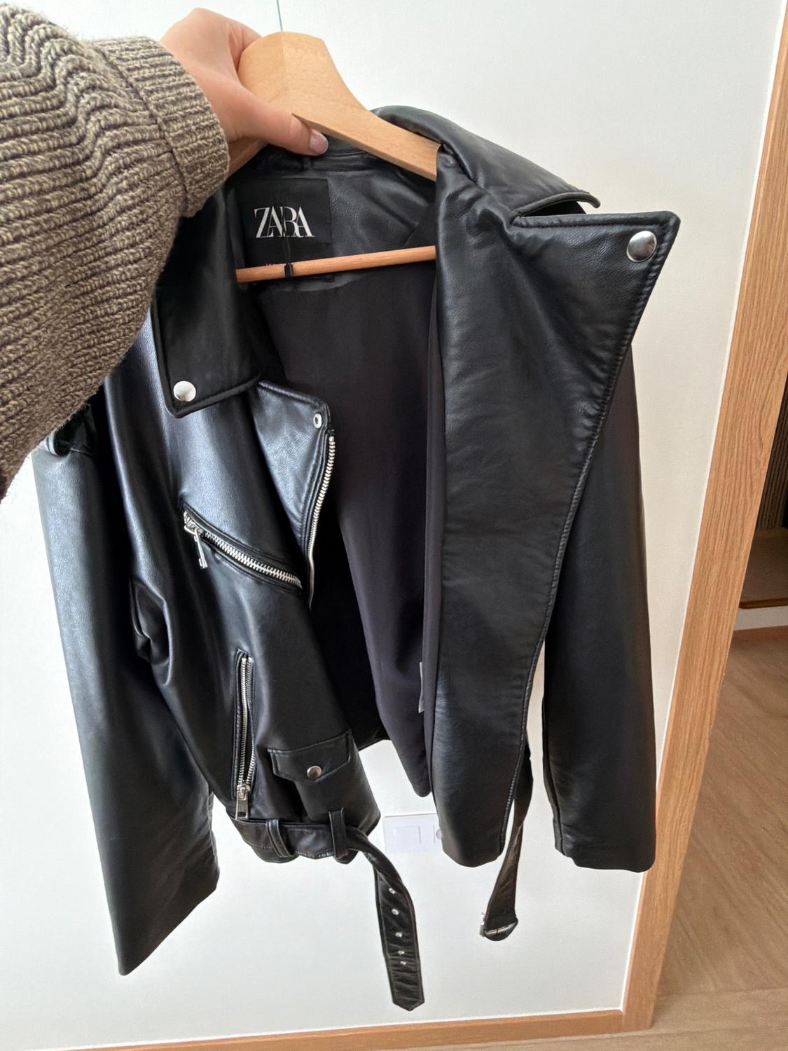 Black leather jacket hanging on a wooden hanger, with zips and a belt. Visible label with the text "ZARA".