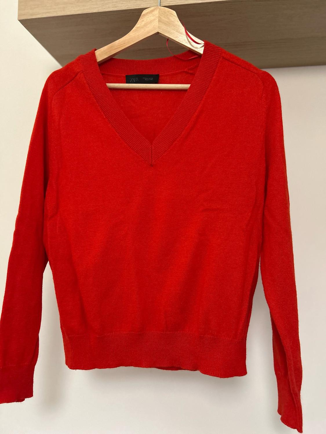 Red knitted jumper, long sleeves and a V-neck, hanging on a wooden hanger in front of a white wall. ZARA label.