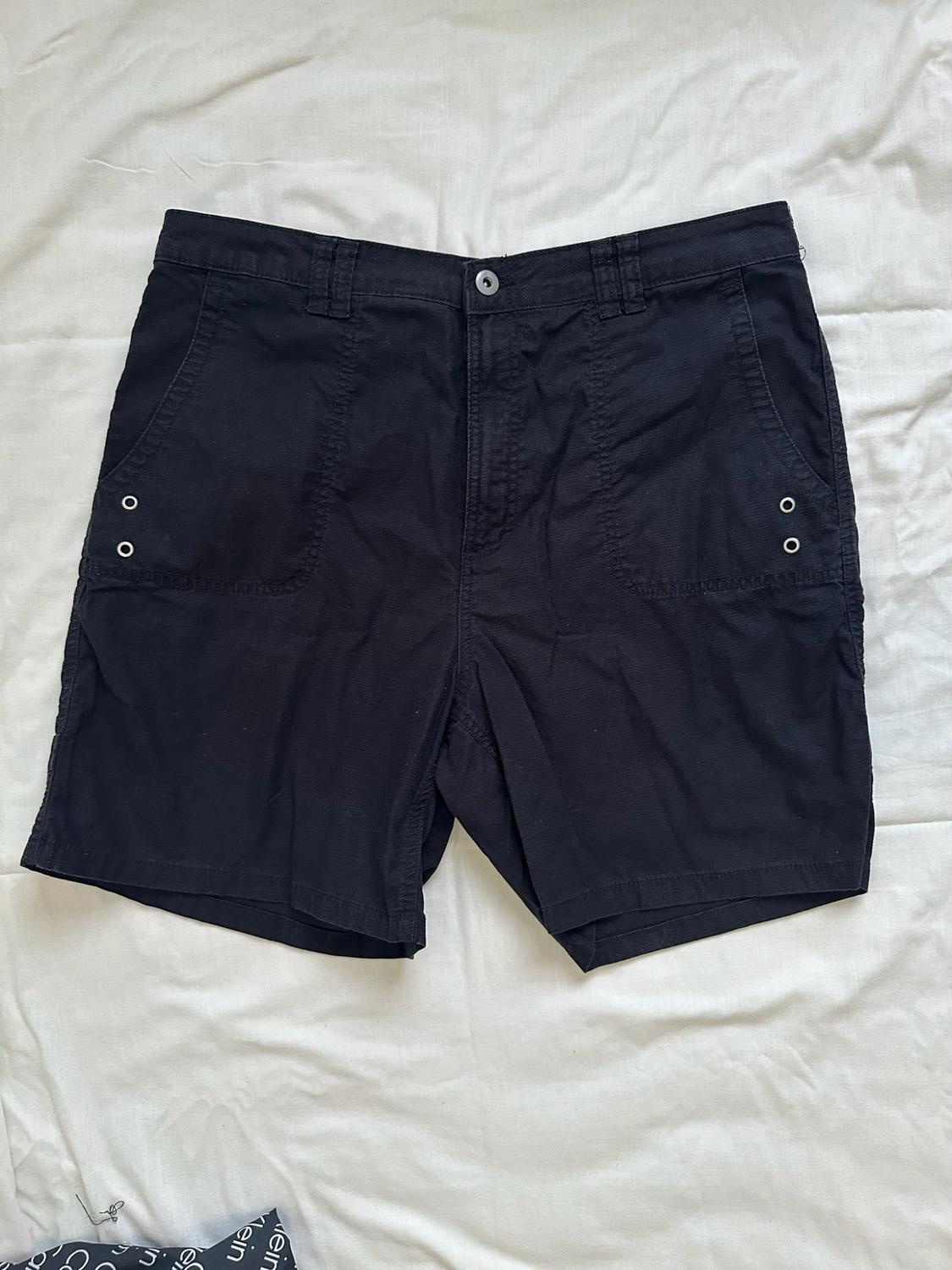 Black shorts with pockets and metallic details, photographed on crumpled white fabric in a relaxed style.