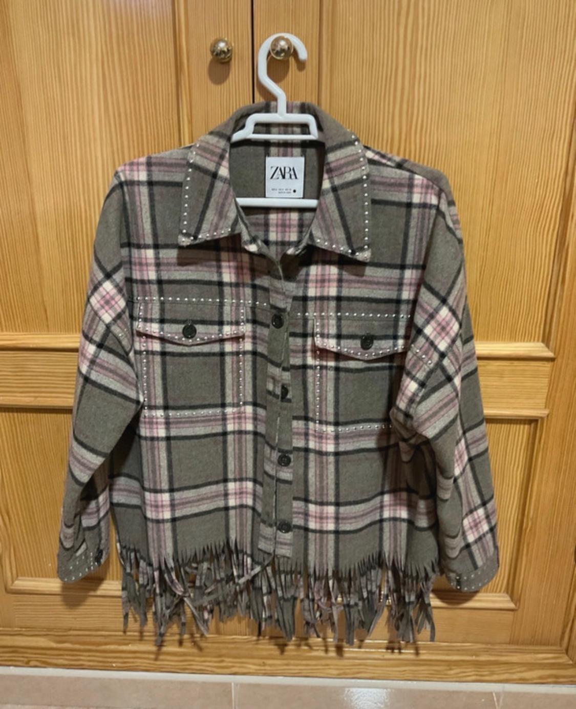 Grey and pink checked jacket with fringing on the hem, hanging on a white hanger in front of a wooden door.