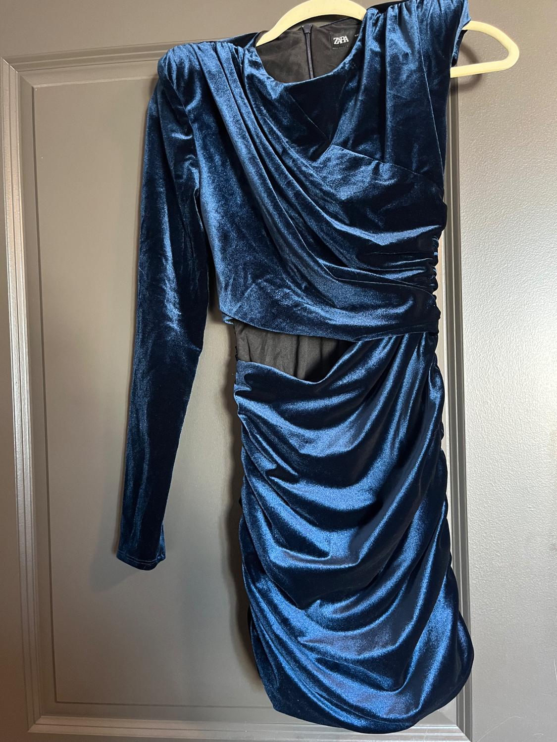 Blue velvet dress with long sleeves and draping, hanging on a hanger over a gray door. Visible tag: Zara.