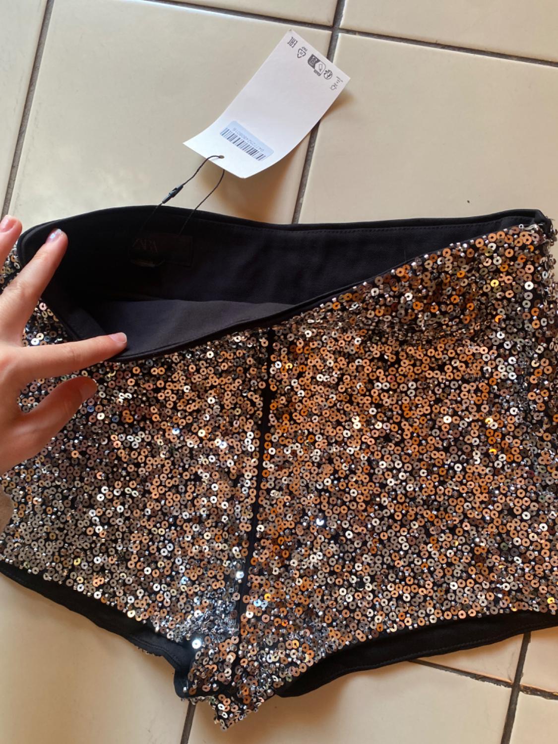 A hand holds up a pair of sequinned shorts over tiled flooring. A white label with the text EAC and the text ZARA is visible on the inside.