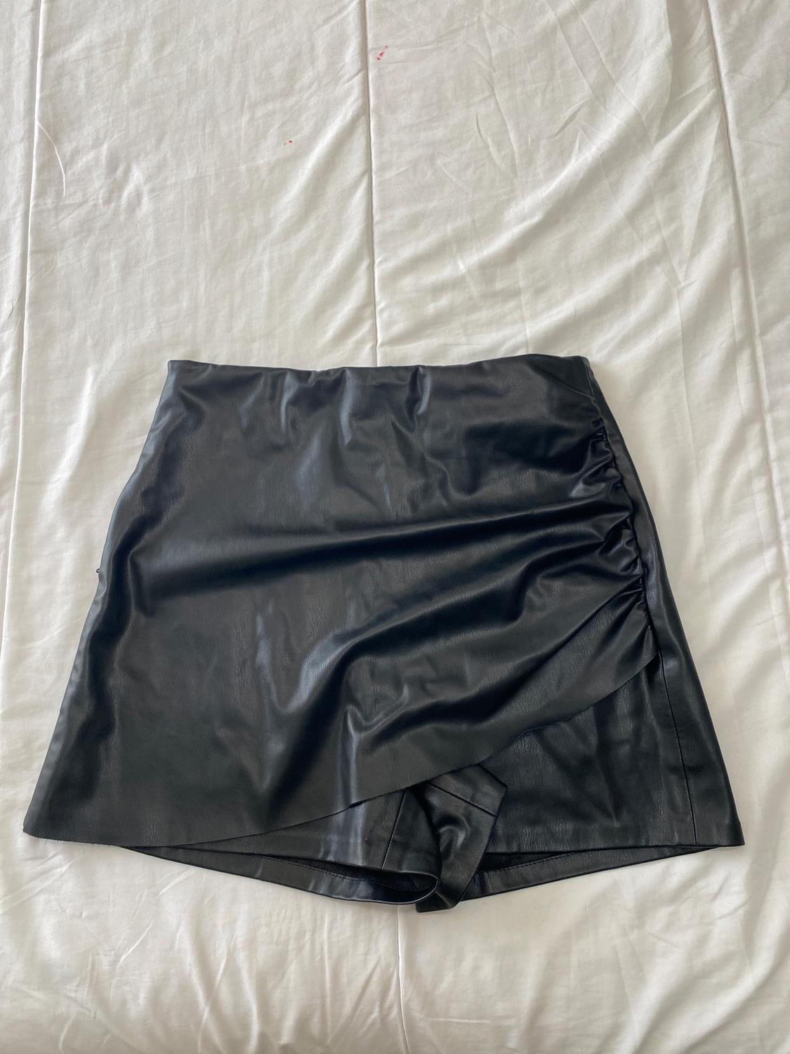 Black, synthetic mini skirt with side pleats, on a wrinkled white surface.