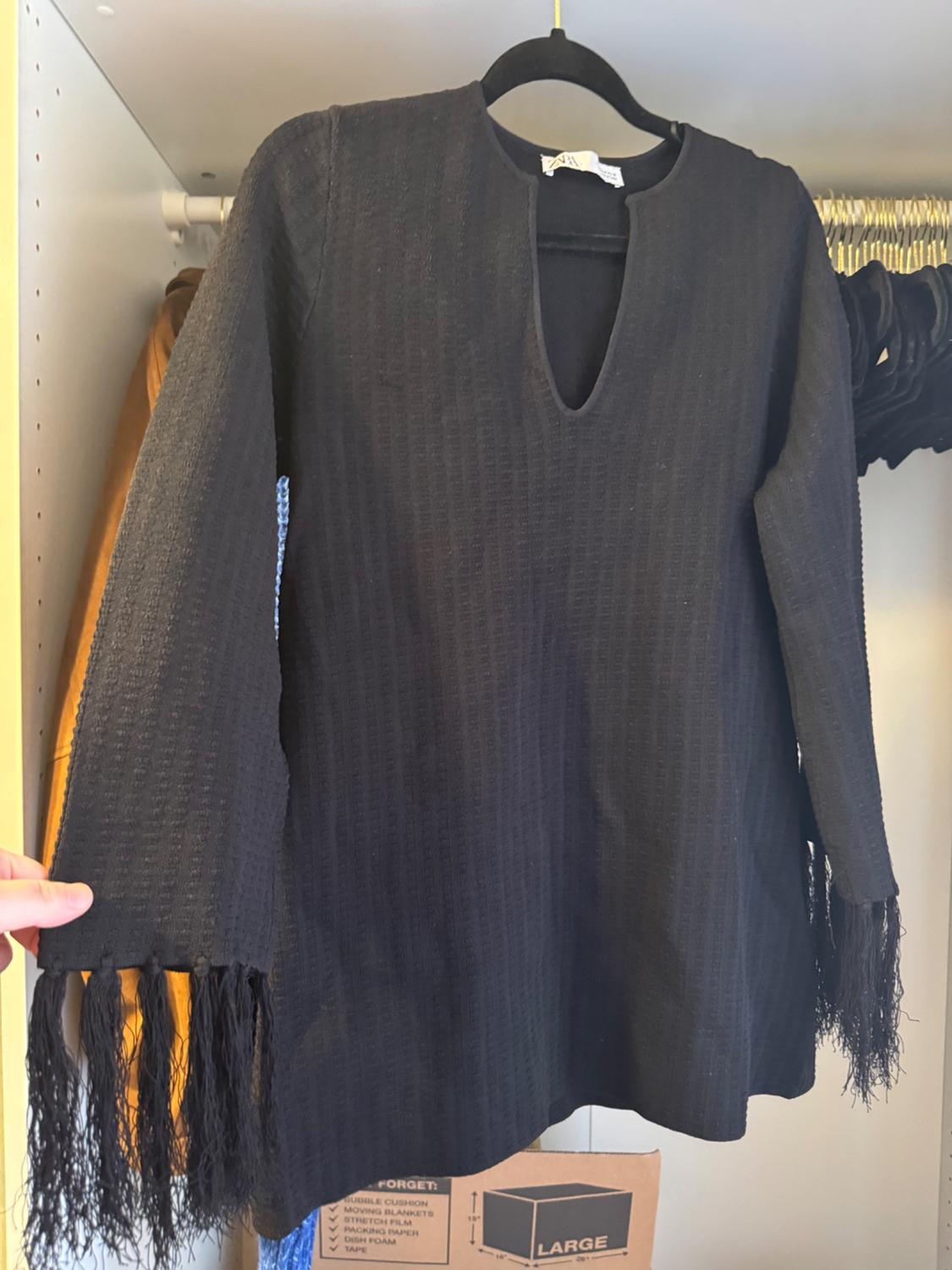 Black long-sleeved blouse with fringes on the sleeves, hanging on a hanger inside a wardrobe. Cardboard boxes in the background.