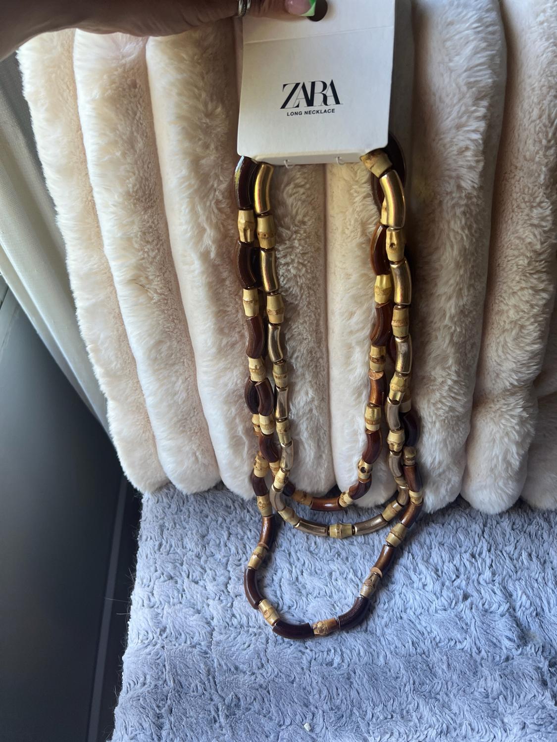 Hand holding a necklace of brown and beige beads against a background of soft textures. Label with the text ZARA LONG NECKLACE.