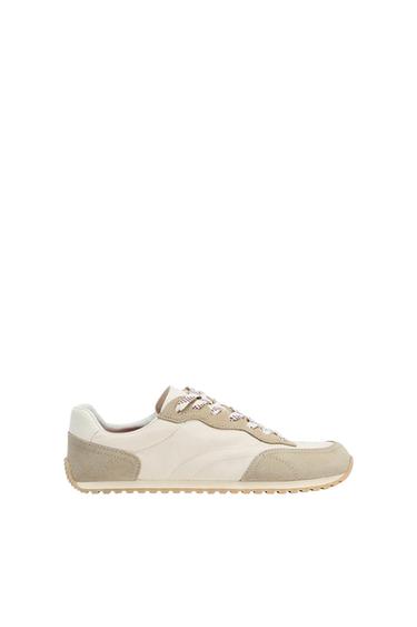 Casual trainers in cream and light beige tones, with laces, textured details and a jagged rubber sole on a white background.