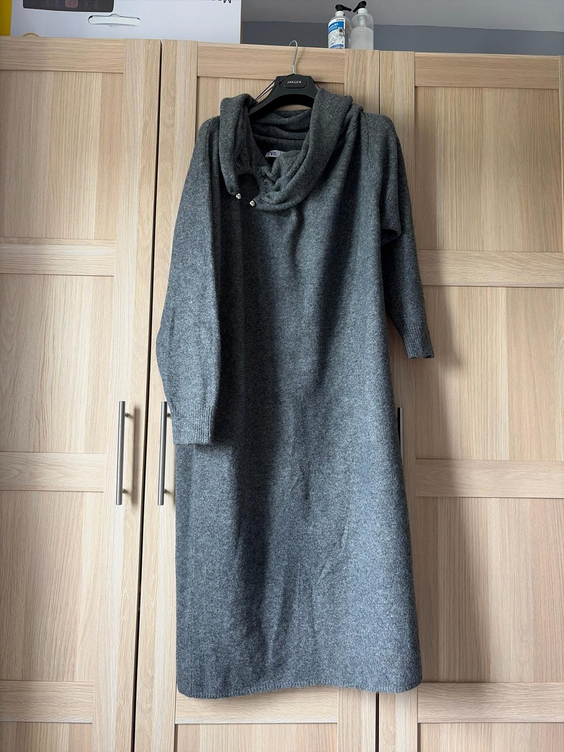 Grey knitted maxi dress with a cowl neck hanging in a wooden wardrobe in a relaxed setting. Text: JAEGER, ZARA.
