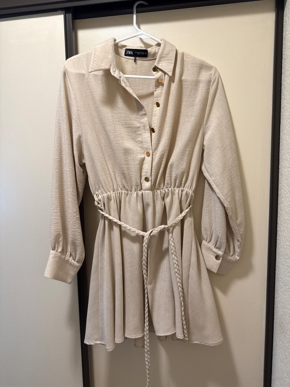 Beige button-down shirt dress with an elastic waist and braided belt, hanging on a door. ZARA text on the label.