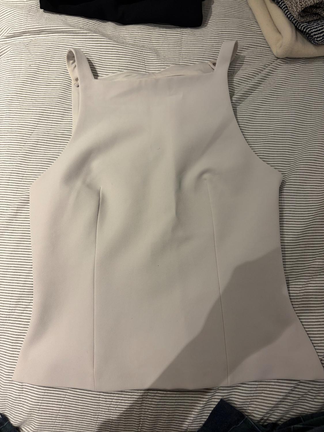 Sleeveless beige top with wide straps on a striped fabric surface. In the background, folded garments in dark and light colours.