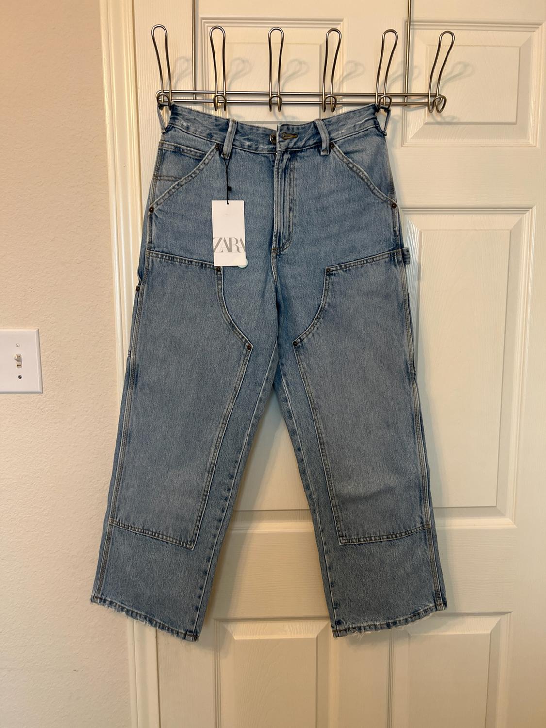 Light blue denim trousers with front panels hanging on a white door. Includes a tag bearing the text ZARA.