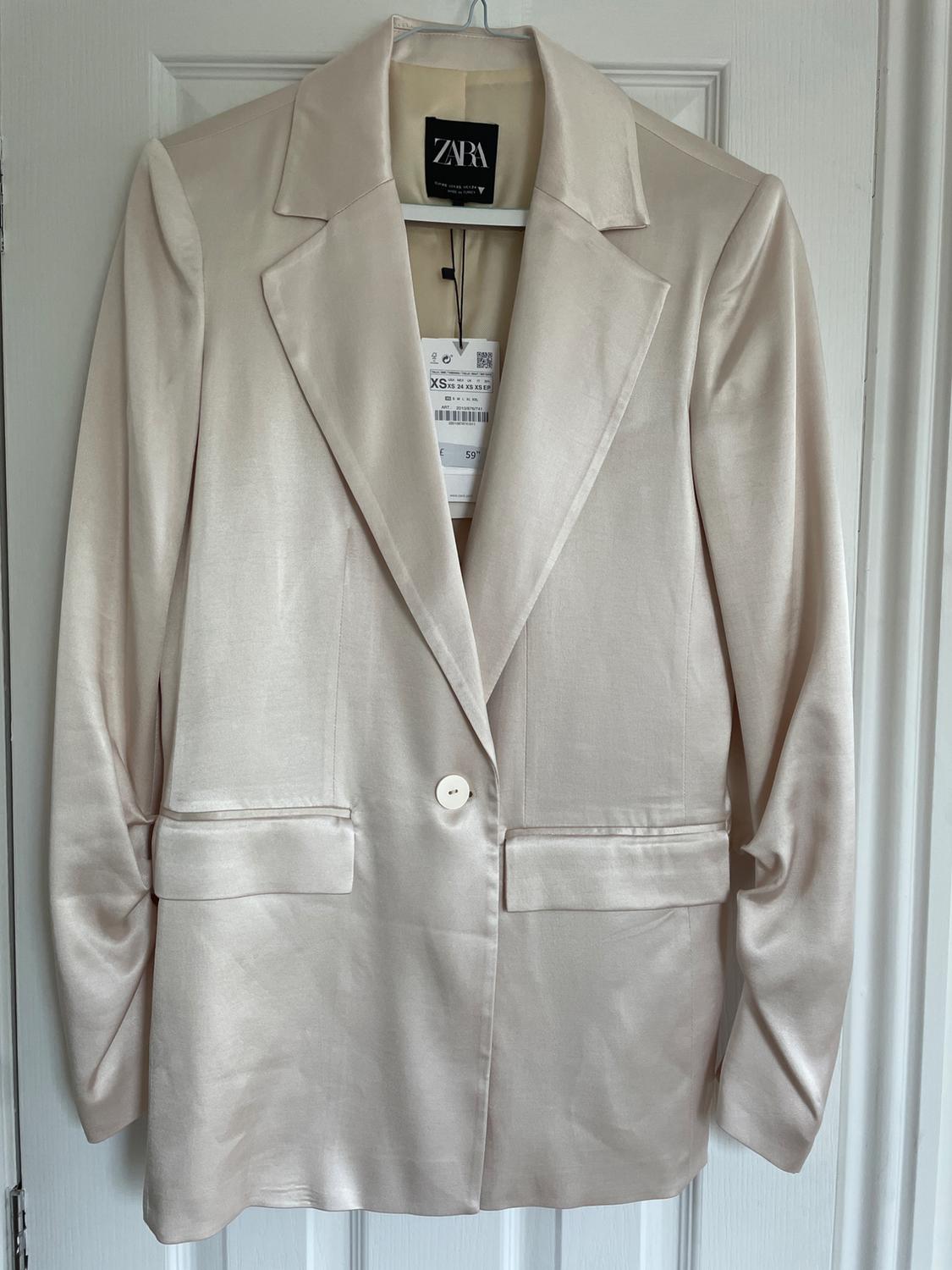 Beige satin jacket hanging on a white door, with Zara tag visible inside and size XS.