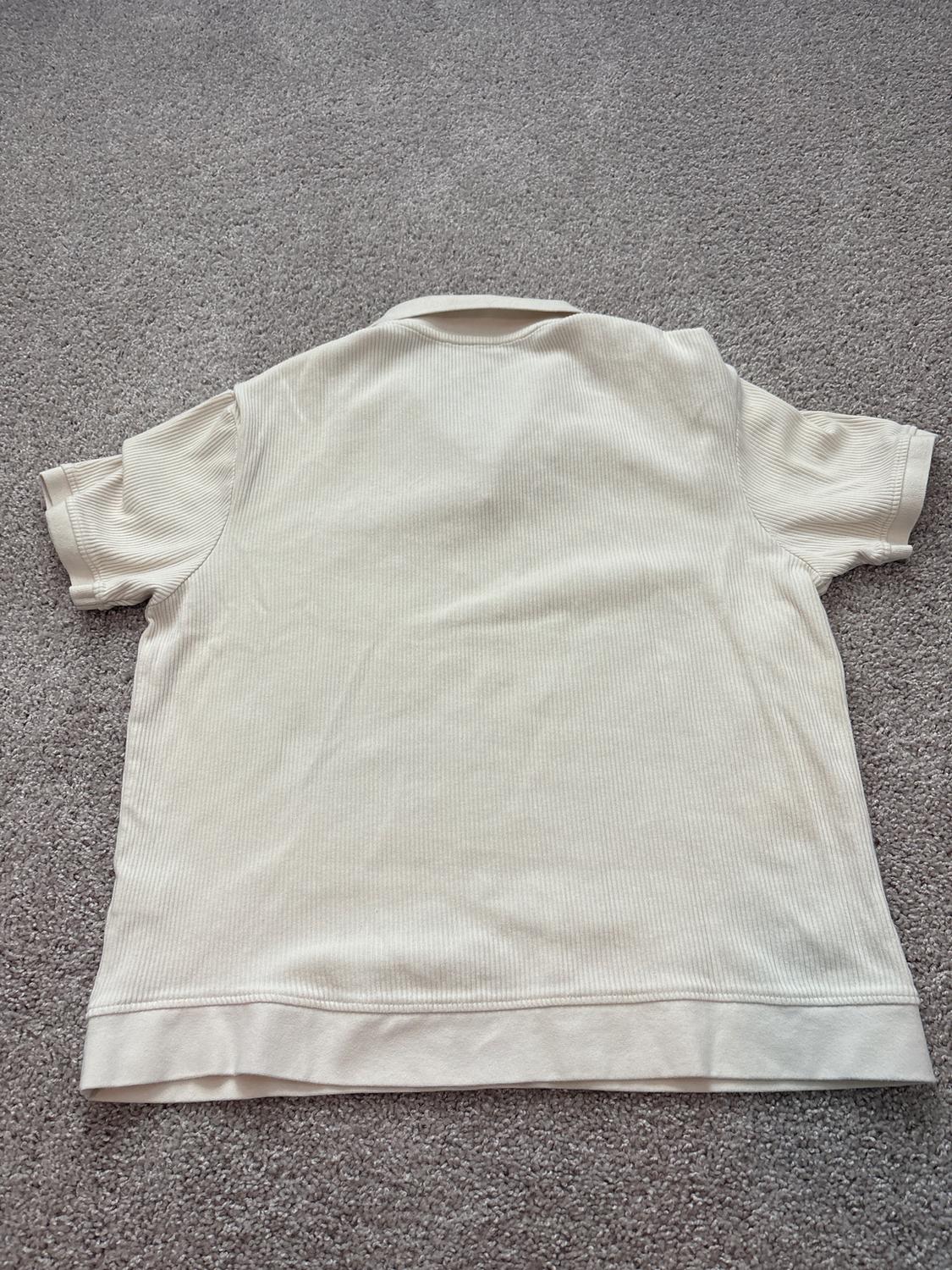 Cream-colored short-sleeved t-shirt seen from the back, on a gray rug. Fabric with a subtly ribbed texture.