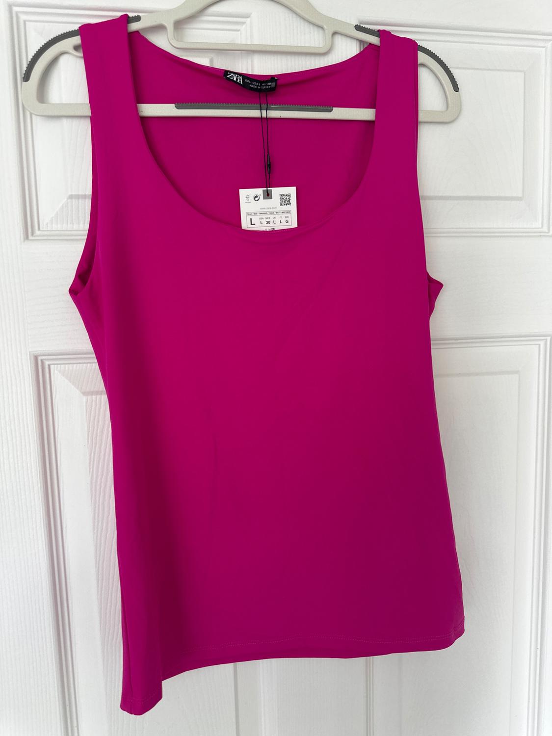 Fuchsia-coloured sleeveless t-shirt hanging on a grey hanger in front of a white door, with visible label including QR code.