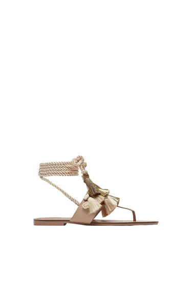 Flat sandal with beige rope straps and gold tassels on a white background. Elegant and summery design.
