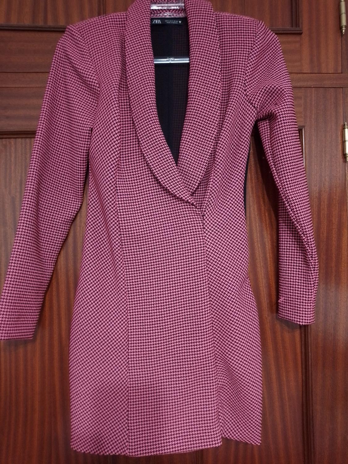 Long coat with small checks in pink and black tones, hanging on a wooden door.