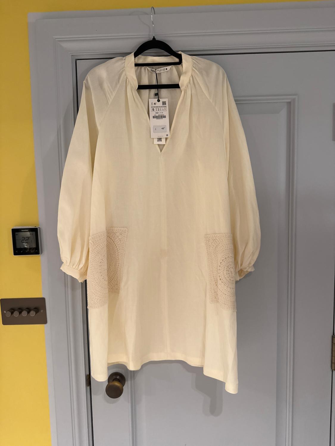 Cream dress with long sleeves and lace details on the pockets, hanging on a grey door. Labels visible on the collar.